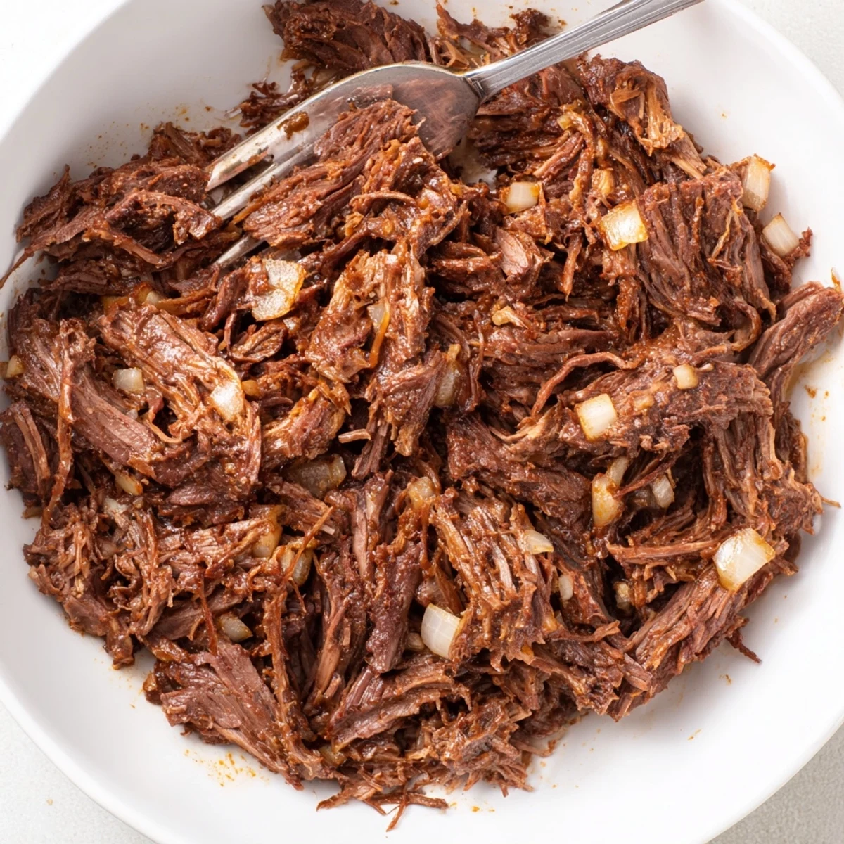Tender slow cooker beef barbacoa shredded in a rich dark red chipotle sauce with fresh white onion pieces scattered on top.